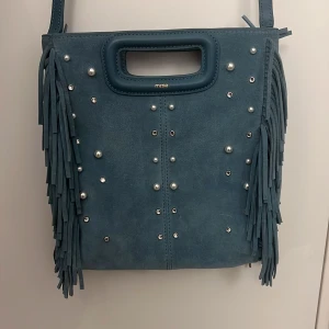 Blå mocka axelväska med fransar från Maje -  ALDRIG ANVÄND (impregnerad)Snygg blå axelväska i mocka från Maje med coola fransar på sidorna och silverfärgade nitar framtill. Väskan har både handtag och lång axelrem. Den finns ej och köpa längre . Köpte för 3,500 ish