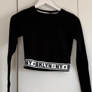 Svart croppad långärmad topp BKLYN - Svart croppad långärmad topp med vit kant längst ner där det står BKLYN NY med svarta stjärnor. Rund halsringning och stretchigt material som känns bekvämt. Perfekt för dig som gillar streetstyle och vill ha en cool look.