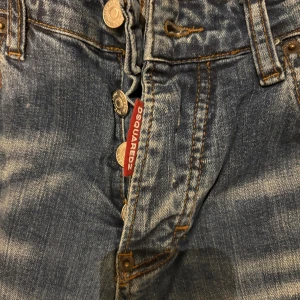 Blå Dsquared2 jeans med patchar - Snygga blå jeans från Dsquared2 med slitningar och coola patchar på framsidan och bakfickan. Jeansen har knappar i metall, klassisk femficksdesign och orangea sömmar. Passformen är slim och materialet är robust denim. Perfekt för dig som gillar streetstyle och detaljer. Detta par har jag användt 5-7 gånger.