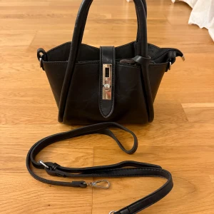 Svart handväska med silverdetaljer - Snygg svart handväska i skinnimitation med stilrena silverdetaljer och praktisk dragkedja. Väskan har både handtag och avtagbar axelrem, samt en dekorativ rem med metallspänne framtill. Perfekt storlek för det viktigaste.