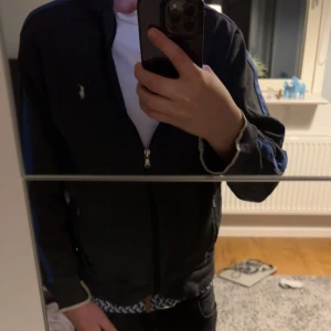 Mörkgrå fullzip från Ralph Lauren  - Mörkgrå fullzip från Polo Ralph Lauren med vit logga på bröstet och blåa detaljer längs ärmarna. Tröjan har dragkedja framtill, två fickor med dragkedja och ribbade muddar. Perfekt för en vardag eller till fina tillfällen. För mer info skicka. Pris kan diskuteras 