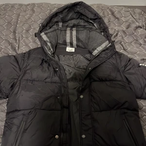 Svart pufferjacka från Burberry - Säljer en svart pufferjacka från Burberry med stor huva och gråmelerat foder med ränder. Jackan har dragkedja, tryckknappar och en tydlig Burberry-patch på ärmen. Perfekt för kalla dagar och riktigt snygg streetstil.