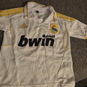 Real Madrid Ronaldo 7 Adidas tröja - Vit Real Madrid fotbollströja från Adidas med gula detaljer och tryck. Framsidan har klubbmärke, Adidas-logga och Bwin-sponsor. Baksidan har Ronaldo och nummer 7 i gult. Tillverkad i lätt och ventilerande ClimaCool-material.