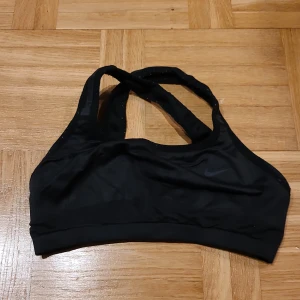 Svart sporttopp från Nike - Vändbar sporttopp. Svart sporttopp från Nike med klassisk logga på framsidan. Modellen har brottarrygg och breda axelband för extra stöd. Materialet är mjukt och stretchigt, perfekt för träning. 