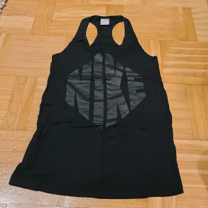 Svart linnetopp från Nike med tryck - Svart linnetopp från Nike med stort, glansigt svart tryck på framsidan. Modellen har brottarrygg och ärmlös design, perfekt för sportiga aktiviteter. Materialet är mjukt och lätt, troligen bomull eller polyester. Snygg och enkel att matcha med träningskläder.