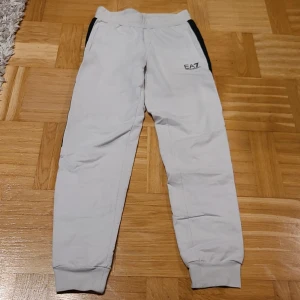 EA7 Emporio Armani ljusgrå mjukisbyxor - Snygga ljusgrå mjukisbyxor från EA7 Emporio Armani med svarta paneler längs sidorna och logga på framsidan. Byxorna har elastisk midja, ribbade muddar vid benslut och är tillverkade i mjukt bomullsmaterial. Perfekta för chill eller träning.