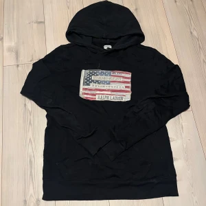 Svart hoodie från Ralph Lauren S - Svart hoodie från Ralph Lauren Denim & Supply med stor patch framtill i amerikansk flaggstil och broderade detaljer. Tröjan har huva, känguruficka och är tillverkad i 100% bomull. Perfekt för dig som gillar streetwear och coola prints. Den har ett litet hål typ där bicepen är men det är verkligen inget man tänker på. Bara att skriva vid frågor. Storleken är S men sitter mer som en M/L