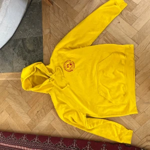 Gul hoodie från Roly med röd print - Gul hoodie från Marabou med stor röd grafisk print på ryggen och smiley-tryck på bröstet. Klassisk känguruficka framtill, snörning i huvan och tryck även på ena ärmen. Perfekt för dig som vill sticka ut med färg och cool design.