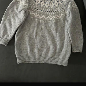 Grå stickad tröja från Lindex - Mysig grå stickad tröja från Lindex med rund hals och lång ärm. Tröjan har ett snyggt mönster i vitt och blått runt halsen som ger en cool nordisk vibe. Perfekt för dig som gillar klassisk stil med en twist.