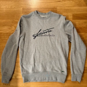 Grå sweatshirt från Lacoste - Grå sweatshirt från Lacoste med klassisk rund hals och lång ärm. Framsidan har ett stort Lacoste-tryck med flera krokodiler och en broderad grön krokodil vid nederkanten. Tröjan är i bomull och har en normal passform som är chill och enkel att bära.