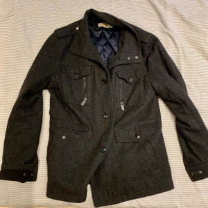 Michael Kors Mörkgrå rock foder - Snygg mörkgrå rock från Michael Kors, med klassisk krage och flera fickor med både knappar och dragkedjor. Insidan har ett quiltat, mörkblått foder som ger extra värme. Perfekt för dig som gillar stilrena och funktionella jackor med detaljer som sticker ut.