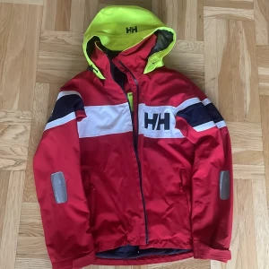 Röd jacka Helly Hansen  - Säljer en röd vindjacka från Helly Hansen med vita och marinblå detaljer samt en neon-gul huva. Den enda skavanken som syns på utsidan är det som syns på tredje bilden. Originalpris är ca 2500kr, så 60% rabatt☺️