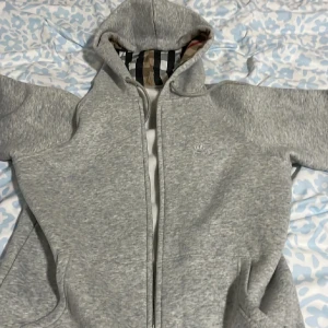 Grå hoodie från Burberry med dragkedja - Säljer en grå hoodie från Burberry med klassiskt rutigt foder i huvan. KOM GÄRNA MED PRISFÖRSLAG