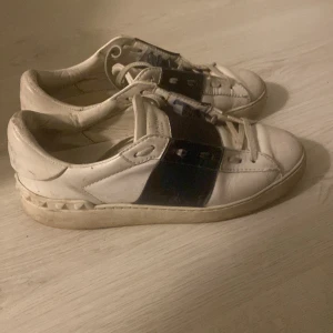 Valentino Garavani Open sneakers vit/svart - Snygga Valentino Garavani Open sneakers i vitt skinn med bred svart rem över vristen och ikoniska nitar på hälen. Klassisk rund tå, platt sula och snörning. Perfekta för dig som vill ha en lyxig och stilren sneaker med edge.