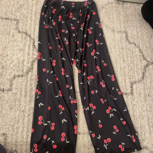 Svarta byxor med körsbärsmönster - Säljer ett par svarta byxor med ett gulligt körsbärsmönster i rött och grönt. Byxorna har en loose passform och breda ben, samt resår i midjan för extra komfort. Perfekta för dig som gillar lekfulla prints och avslappnad stil.