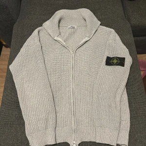 Grå stickad kofta från Stone Island - Säljer en grå stickad kofta från Stone Island med dragkedja och hög krage. Klassisk patch med logga på vänster ärm. Koftan har långärmad passform och grovstickad struktur som ger en schysst vibe. Perfekt för dig som gillar streetwear och snygga detaljer.passar bra nu till hösten, priset kan alltid diskuteras via snabba affärer 