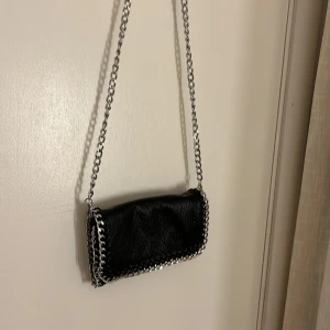 Svart väska  - Snygg och stilren svart väska i mjukt skinnimitation med silverfärgade kedjedetaljer runt kanterna. Väskan har en avtagbar axelrem i metallkedja, vilket gör att den kan bäras både som axelväska eller clutch. 
