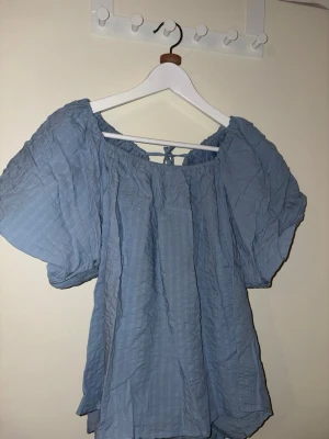 Blå blus med puffärm från Lindex - Supersöt ljusblå blus från Lindex med puffiga korta ärmar och knyt i ryggen. Tillverkad i ett mjukt bomullstyg med diskret rutstruktur. Perfekt till jeans eller kjol för en chill look. Prislapp fortfarande på