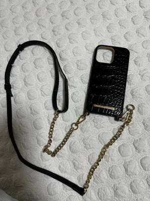 Ideal of Sweden mobilskal med kedja - Elegant svart mobilskal till IPhone 14 pro max med krokodilstruktur och guldfärgad kedja/axelrem från Ideal of Sweden. Passar större iPhone-modeller. Skalet är i fint skick utan synligt slitage och ger både skydd och stil till din mobil.