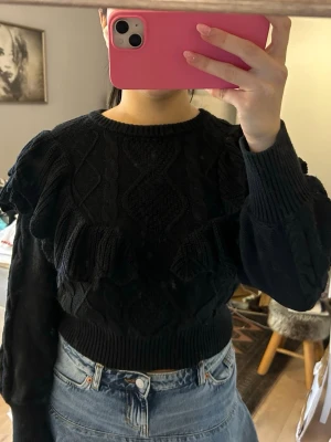Svart stickad tröja med volanger - Säljer en svart stickad tröja med volangdetaljer och kabelstickat mönster. Tröjan har rund halsringning, långa ärmar och ribbade muddar. Perfekt för dig som vill ha en trendig och mysig look till hösten eller vintern.