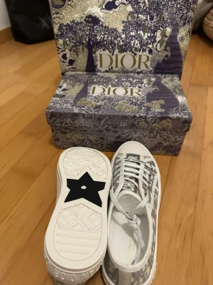 Dior sneakers med monogrammönster - Säljer ett par snygga Dior sneakers i vitt och grått med klassiskt Dior-monogram i tyg. Skorna har vita snören, rund tå och platt sula med en svart stjärndetalj undertill. Märket syns tydligt på plösen och sidan. Perfekta för dig som vill ha något exklusivt och trendigt.