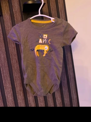 Grön body med tryck storlek 62 - Söt grön body med korta ärmar och tryck framtill av en gul sengångare och texten 'no panic'. Bodyn har gul kant på insidan av halsen och knäppning på axeln och i grenen. Perfekt för chill vibes och lek. Storlek 62