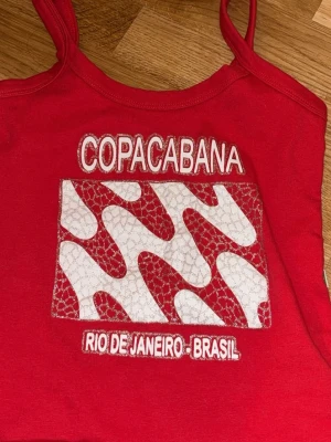 Röd Copacabana linnetopp - Snygg röd linnetopp med smala axelband och broderad text 'Copacabana' samt 'Rio de Janeiro - Brasil' framtill. Toppen har ett vågigt mönster i vitt och rött med glittriga detaljer. Perfekt för sommaren. Den är köpt i Brasilien. Passar både xs och S, säljer den eftersom den aldrig kom till användning.