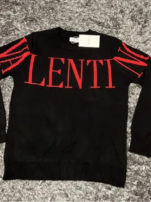 Svart tröja med röd VALENTINO-text - Snygg svart tröja från Valentino med stora röda bokstäver över bröstet och armarna. Tillverkad i 100% bomull och har långa ärmar samt ribbade muddar. Perfekt för dig som vill sticka ut med en exklusiv streetwear-look.