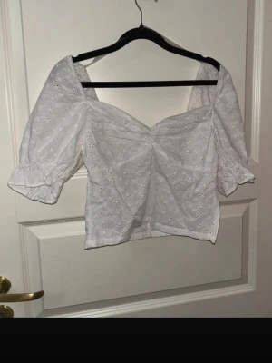 Vit blus med broderade detaljer - Söt vit blus med puffärmar och v-ringad front. Blusen har broderade blommönster över hela tyget och en dold dragkedja i sidan. Tillverkad i bomull för en luftig känsla, perfekt för varma dagar. Ärmarna har resår vid avslut för en fin siluett.