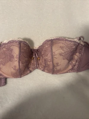 Lila spets-bh från Masquerade - Säljer en lila bh från Masquerade, modell Antoinette. Bh:n har vacker spets med blommigt mönster, liten rosett framtill och dekorativa detaljer. Axelbanden är justerbara och kuporna ger ett fint lyft. Perfekt för dig som gillar feminina detaljer.