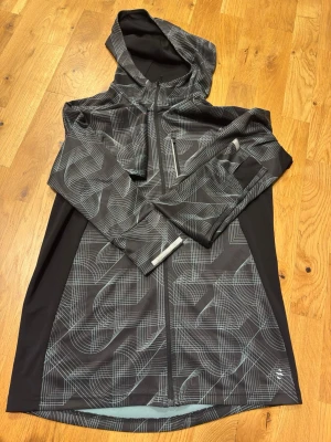 Svart vindjacka med mönster från H&M - Säljer en svart vindjacka från H&M Sport med grått grafiskt mönster. Jackan har huva, hel dragkedja framtill och en bröstficka med dragkedja. Tillverkad i lätt polyester, perfekt för löprundan eller promenaden. Snygg och sportig look!