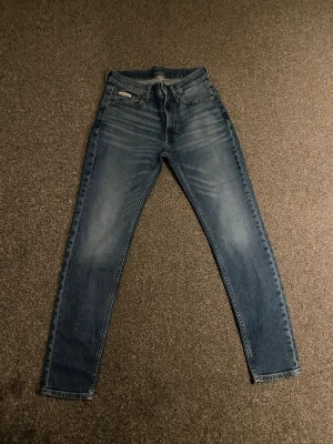Calvin Klein Slim Taper jeans blå - Säljer ett par blå Calvin Klein jeans i modellen Slim Taper. Jeansen har klassisk femficksdesign, normal midja och smalare ben. Tillverkade i slitstarkt bomullsmaterial med snygg tvättad look och diskreta CK-detaljer på knappar och ficka.