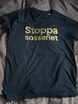 Mörkblå t shirt med märke på - Mörkblå T-shirt i bomull med rund hals och korta ärmar. Framsidan har ett stort tryck i guld med texten 'Stoppa sosseriet' som verkligen sticker ut. Perfekt för dig som vill göra ett statement.