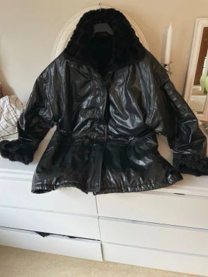 Svart pufferjacka med fuskpäls - Säljer en svart pufferjacka med glansig yta och mjukt fuskpälsfoder. Jackan har dragkedja framtill, stora fickor med dragkedja och knappar samt bred krage i fuskpäls. Perfekt för kalla dagar och riktigt snygg oversized look.