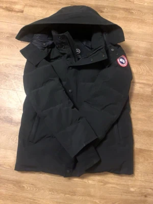 Svart dunjacka från Canada Goose - Säljer en svart dunjacka från Canada Goose med huva och klassisk logga på ärmen. Jackan har dragkedja och knappar framtill, samt flera fickor. Perfekt för kalla vinterdagar och riktigt varm tack vare dunfyllningen.