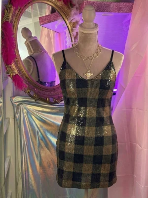 Glittrig rutig kortklänning made in italie - Supersnygg kort klänning med tunna axelband. Klänningen har ett rutigt mönster i svart och guld, täckt av glittriga paljetter. Materialet är polyester och den har dragkedja i sidan. Perfekt för dig som vill sticka ut.