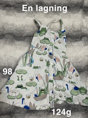 Söt klänning med grodmönster, stl 98 - Supergullig klänning med knappar framtill och axelband. Klänningen är vit med färgglatt mönster av grodor, näckrosor, bin och blommor. Perfekt för varma dagar och lek. 🌸 Finns en lagning. Storlek 98