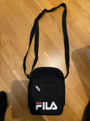 Svart axelväska från FILA - Säljer en svart axelväska från FILA med justerbar rem och dragkedja framtill. Väskan har en enkel, stilren design med FILA-logga i vitt och rött på framsidan. Perfekt storlek för det viktigaste och tillverkad i slitstark polyester.