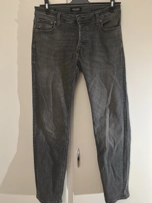 Grå jeans från Jack & Jones - Säljer ett par grå jeans från Jack & Jones med klassisk femficksdesign och raka ben. Jeansen har en snygg tvättad look och är tillverkade i slitstarkt denimtyg. Passar perfekt till en avslappnad stil och är lätta att matcha med olika plagg.