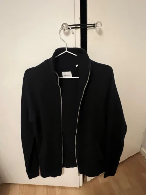 Svart stickad kofta från Jack & Jones - Säljer en svart stickad kofta från Jack & Jones med dragkedja framtill och hög krage. Perfekt för lager på lager och enkel att matcha med olika outfits. Klassisk och stilren design med lång ärm.