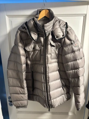 Grå Moncler dunjacka med huva - Säljer en grå Moncler dunjacka med huva och dragkedja. Jackan har två bröstfickor med knappar, broderad Moncler-logga på ärmen och quiltad design. Perfekt för kalla dagar och riktigt snygg till streetwear-stilen.