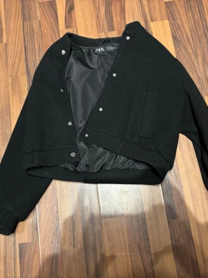 Svart bomberjacka från Zara - Säljer en svart bomberjacka från Zara med ribbade muddar och tryckknappar framtill. Jackan har en croppad passform och är tillverkad i ullblandning med slät insida. Enkel och stilren design som passar till många olika looks.