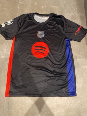FC Barcelona Lamine Yamal tröja - Säljer en svart FC Barcelona matchtröja med röda och blå detaljer, Spotify-logga på bröstet och Lamine Yamal samt nummer 10 på ryggen. Tröjan har officiella klubbmärken och Champions League-märke på ärmen. Perfekt för dig som älskar fotboll och Barca.