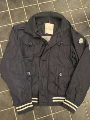 Mörkblå Windbreaker Moncler - Snygg mörkblå bomberjacka från Moncler med klassisk logga på ärmen, fyra stora fickor framtill och ribbade muddar med vita ränder. Jackan har en mjuk grå insida och stängs med både dragkedja och knappar. Perfekt för dig som vill ha en stilren och sportig look.