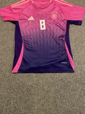 Adidas Tyskland Kroos tröja rosa/lila - Säljer en Adidas Tyskland landslagströja med Kroos och nummer 8 på ryggen. Tröjan har en snygg gradient från rosa till lila med geometriskt mönster, vita detaljer och fyra stjärnor på bröstet. Tillverkad i polyester med Aeroready-teknologi.