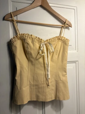 Gul korsettopp med rosett från Zara - Supersöt ljusgul korsettopp från Zara med smala axelband och rynkad kant upptill. Även justerbara axelband! Toppen har en dekorativ vit rosett framtill och markerade sömmar som ger en snygg siluett. Perfekt för sommarens alla tillfällen! Säljer då den aldrig kommit till användning!