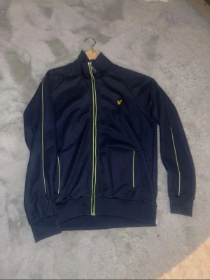 Marinblå zip hoodie från Lyle & Scott - Säljer en marinblå hoodie från Lyle & Scott i storlek L. Tröjan har hel dragkedja framtill, gul/grön detaljer längs dragkedjan och ärmarna samt Lyle & Scott-logga på bröstet.