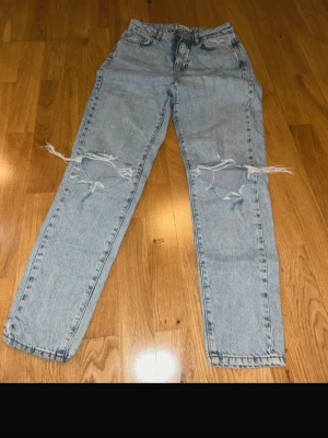 Ljusblå ripped mom jeans - Säljer ett par ljusblå mom jeans från Gina Tricot med stora slitningar över båda knäna. Klassisk femficksmodell, hög midja och raka ben. Jeansen är i ett mjukt bomullsmaterial och har dragkedja och knapp framtill.