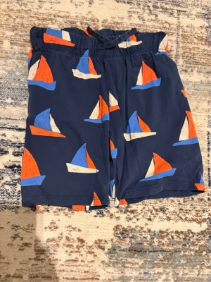 Blå shorts med segelbåtar från H&M - Säljer ett par mörkblå shorts från H&M med färgglatt segelbåtsmönster i orange, blått och vitt. Shortsen har resår i midjan och snörning för extra komfort. Perfekta för sommardagar och strandhäng.