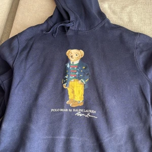 Mörkblå hoodie från Ralph Lauren - Säljer en snygg mörkblå hoodie från Ralph Lauren med den ikoniska Polo Bear på framsidan. Tröjan har en klassisk design med huva. Perfekt för en avslappnad stil. Är stl L 